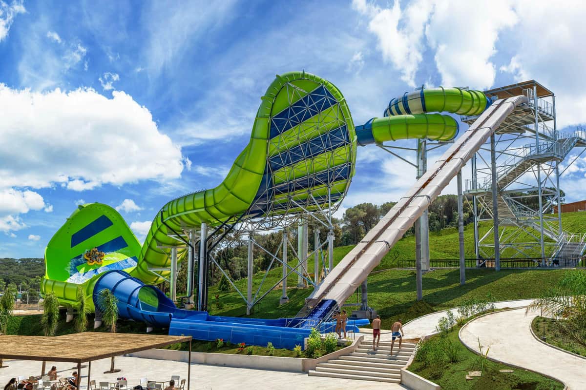 A large green and blue water slide structure with people walking nearby under a sunny sky.