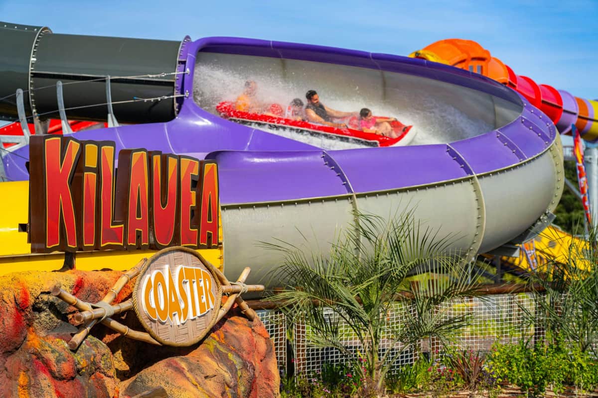 People riding a water slide called the ‘Kilauea Coaster’, featuring a purple tube, colourful signage and tropical plants in the background.