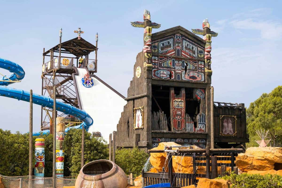 A water park slide near a themed wooden structure with totem poles and colorful designs, surrounded by rocks and greenery.