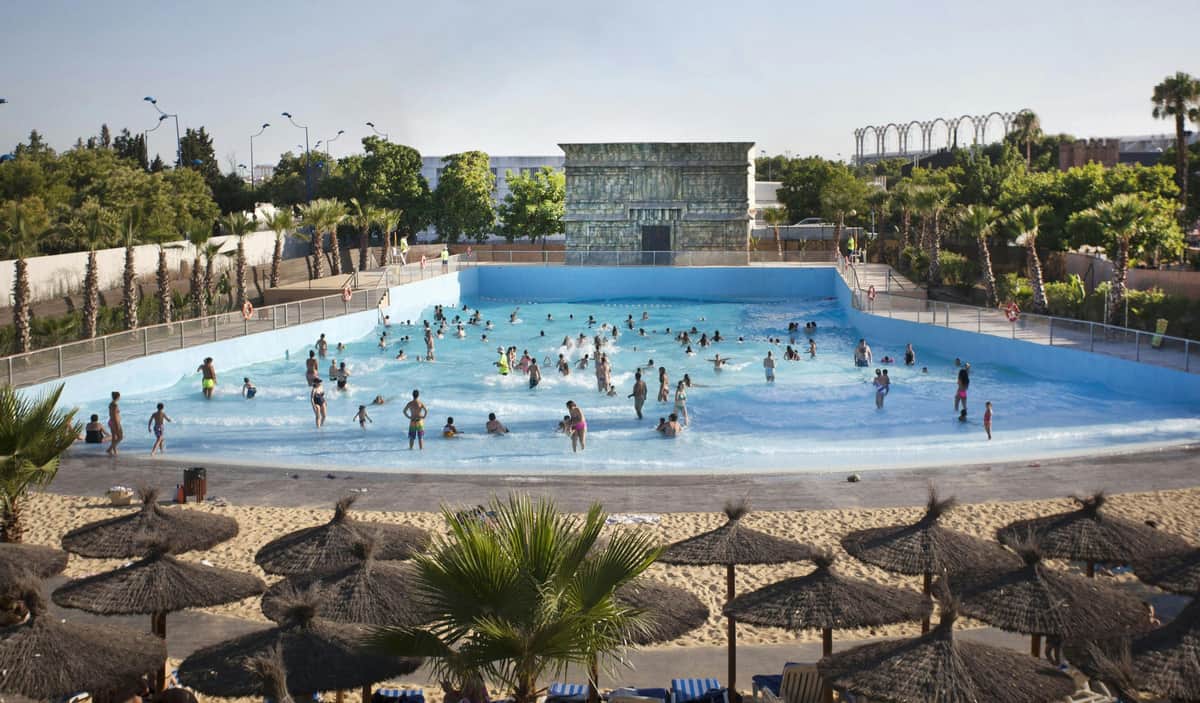 People enjoying a wave pool at a water park, with straw umbrellas in the foreground and a decorative structure in the background.