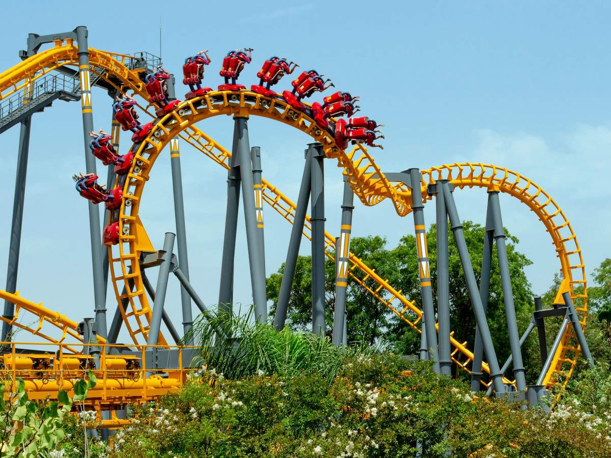 People riding a yellow and red rollercoaster with loops and turns, surrounded by greenery and a clear blue sky.