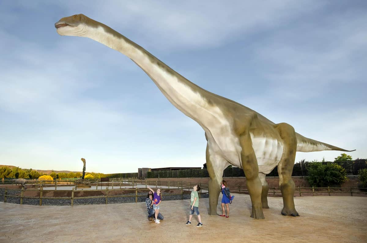 Four people standing near a large dinosaur statue in an outdoor setting with a clear sky in the background.
