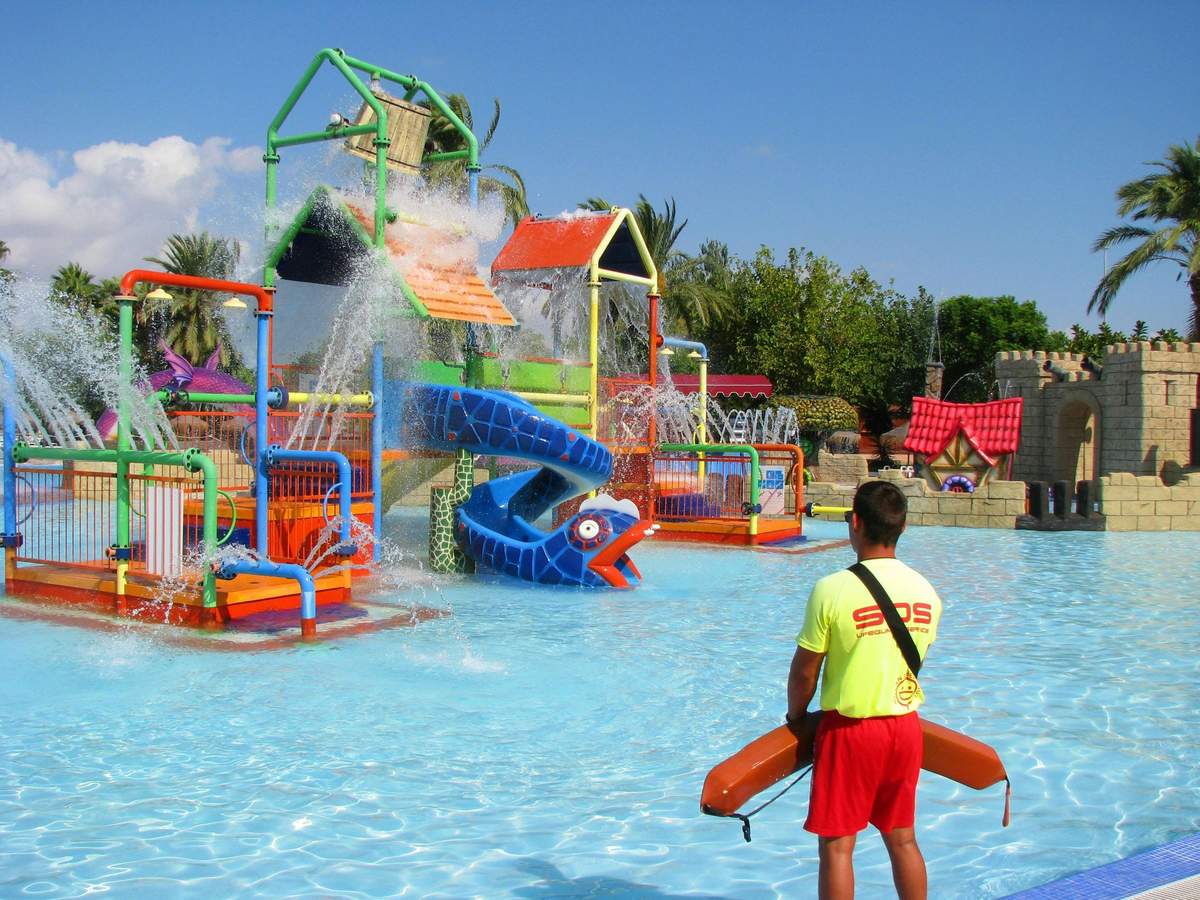 A lifeguard in a bright yellow shirt watches over a colorful water play structure with slides and water sprays in a pool.