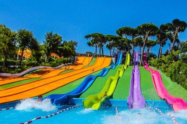 Colourful water slides with people sliding into a pool, surrounded by trees and greenery under a clear blue sky.