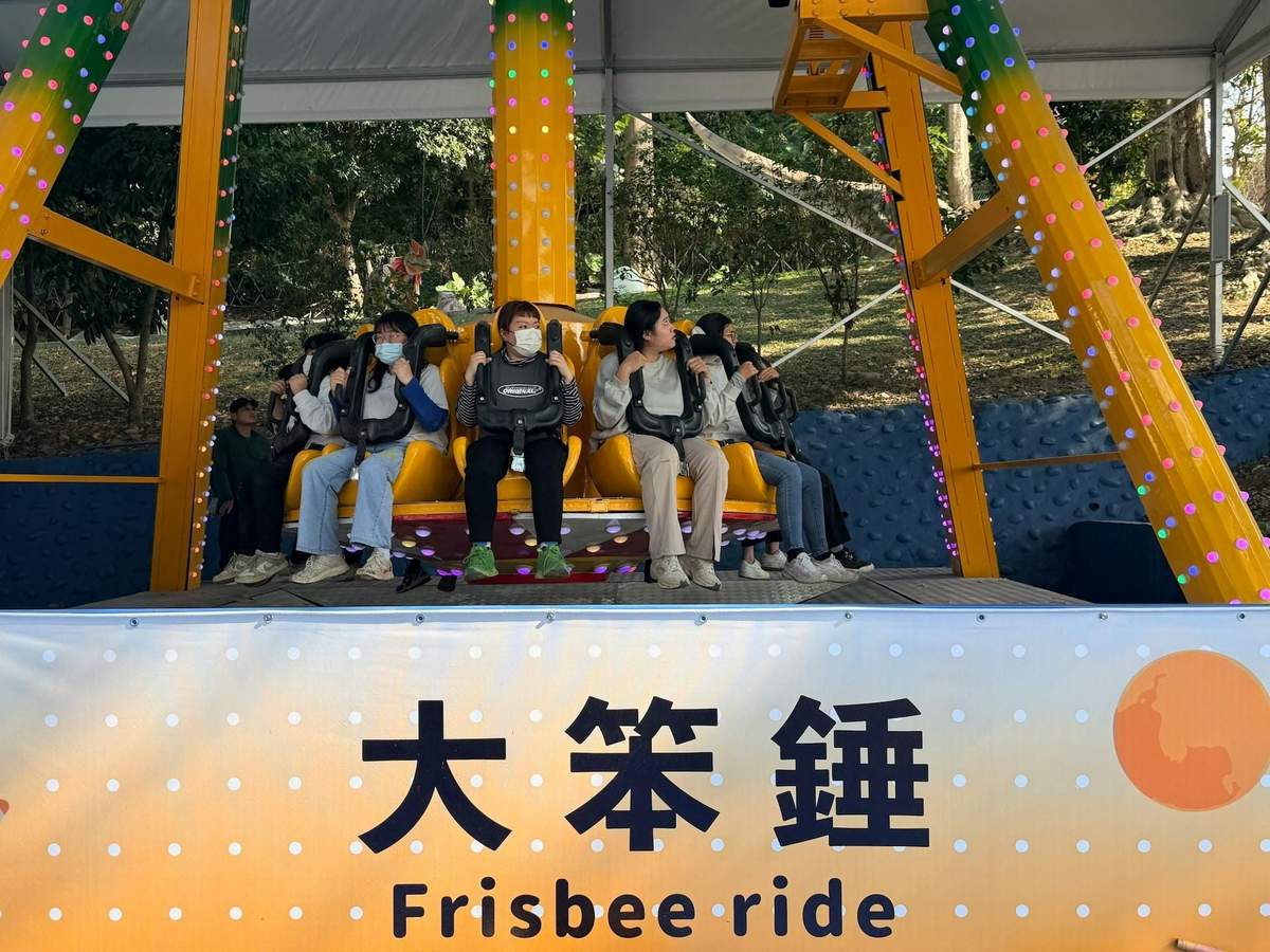 A group of people seated on an amusement ride named 'Frisbee ride,' with safety restraints, under a canopy in a park.
