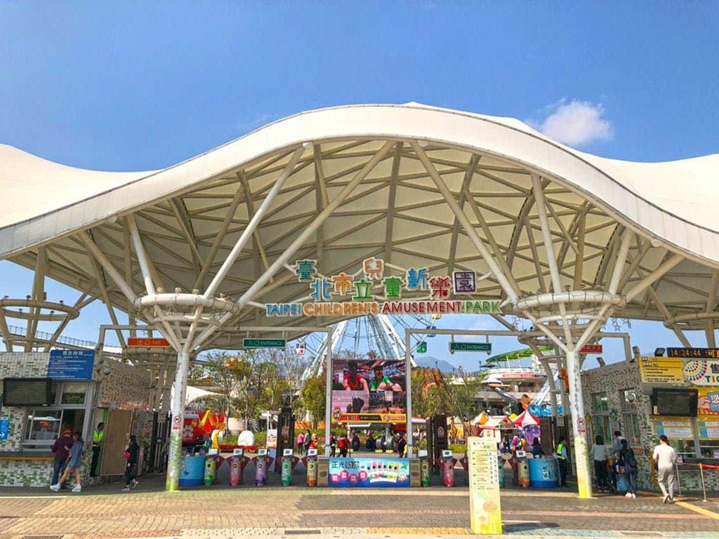 A colorful children's train ride near a ferris wheel and concession stand in a sunny amusement park setting.