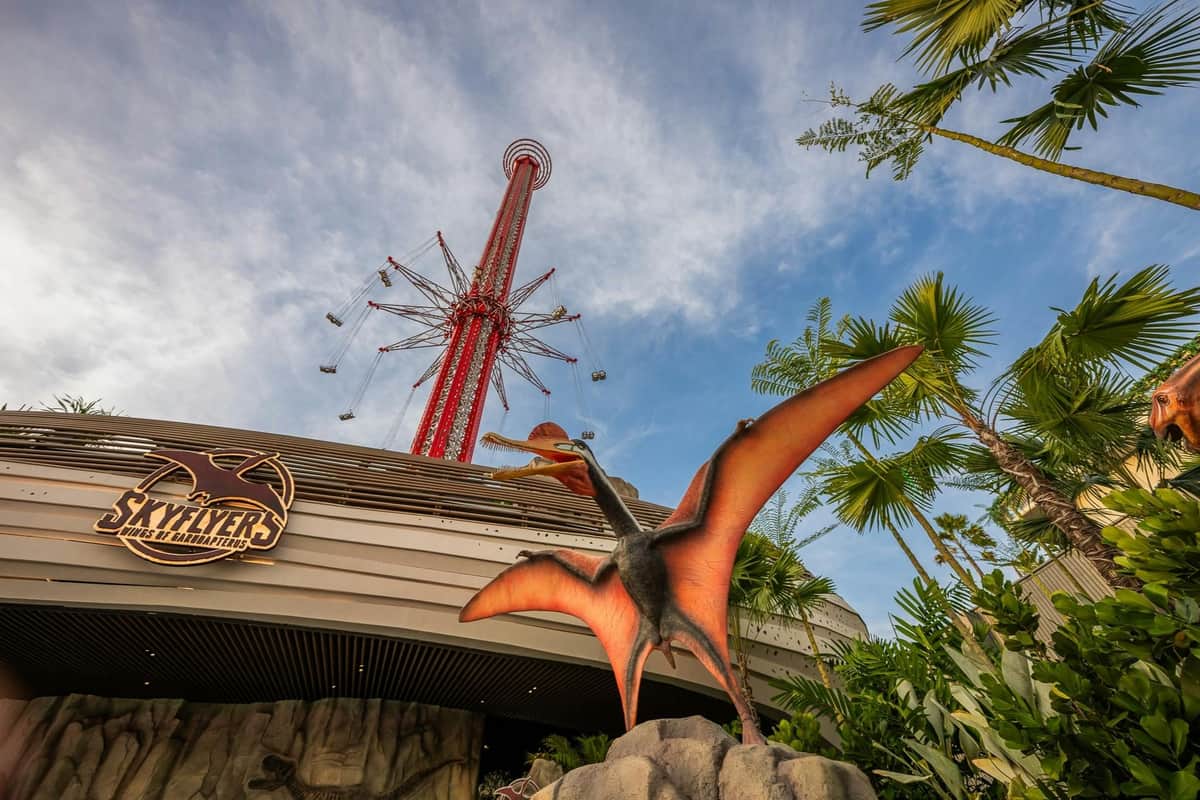 A statue of a flying dinosaur with orange wings is in front of an amusement park entrance, with a tall red ride visible in the background.