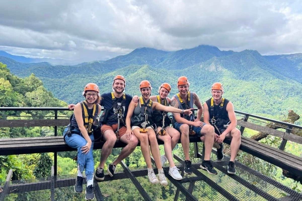 Five people wearing helmets and harnesses sit on a platform with lush green mountains and a cloudy sky in the background.