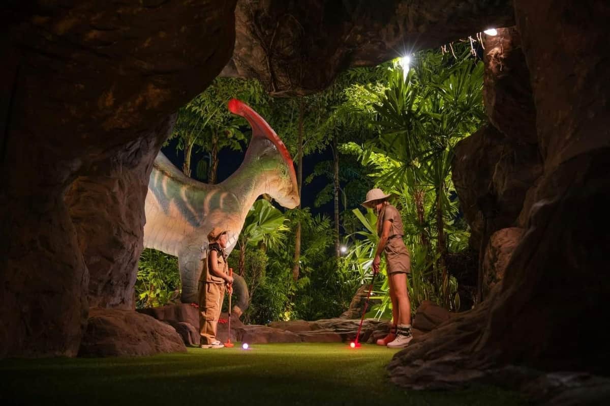 Two people playing mini-golf in a jungle-themed setting with a large dinosaur statue and lush greenery around them.