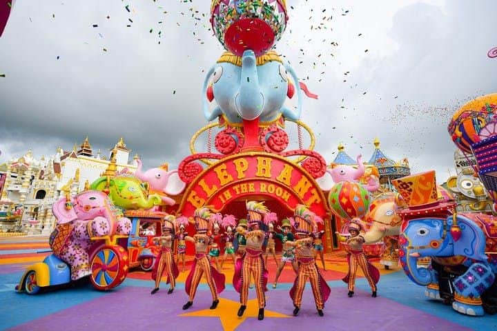 Performers in colorful costumes dance in front of a vibrant "Elephant in the Room" carnival display. Confetti falls from above.