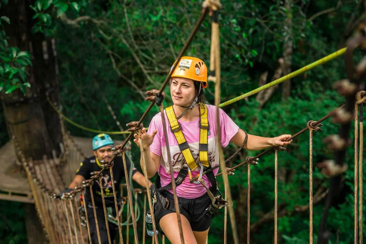Two people wearing helmets and harnesses navigate a rope bridge in a lush forest setting.