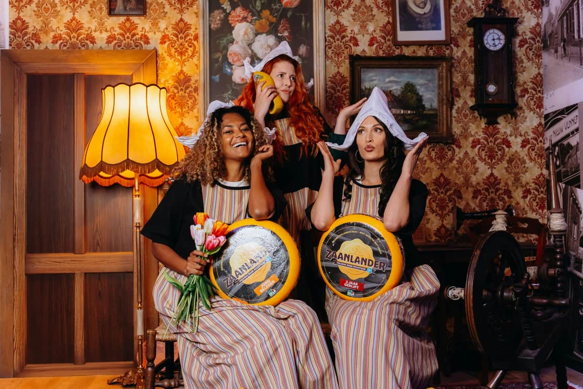 Three women in traditional attire holding wheels of cheese and tulips, posing in a vintage-themed room with floral wallpaper.