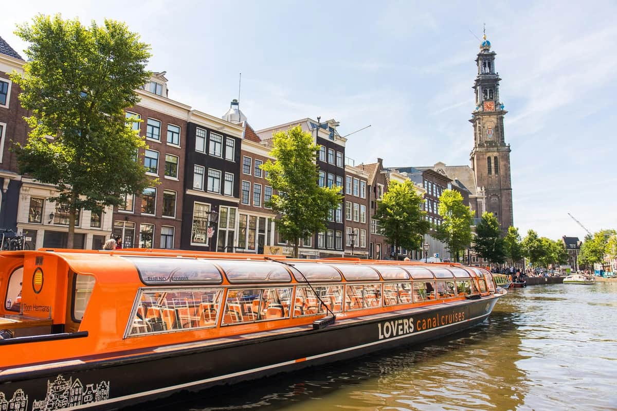 Orange canal cruise boat docked in an Amsterdam canal, with historic buildings and a church tower in the background.