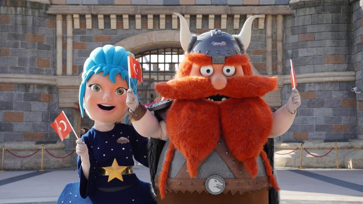 Two costumed characters, one with blue hair and the other dressed as a viking, hold Turkish flags in front of a stone building.