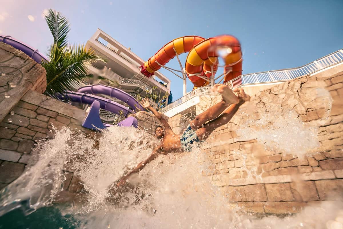 A person splashing into a pool from a slide, with colorful water slides and a building in the background under a clear sky.