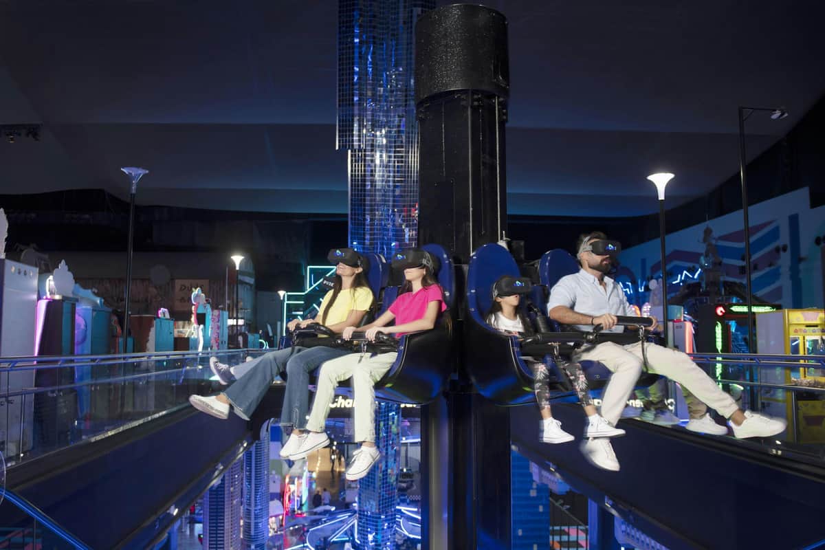 Dubai Play DXB Four people wearing VR headsets are seated on a ride inside a futuristic, neon-lit indoor amusement park.
