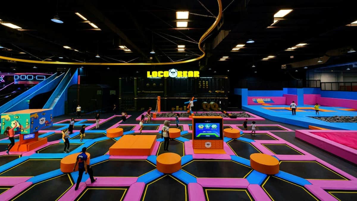 Indoor trampoline park with colorful mats, platforms, and people jumping. A sign reads 'Loco Bear'. Brightly lit area.