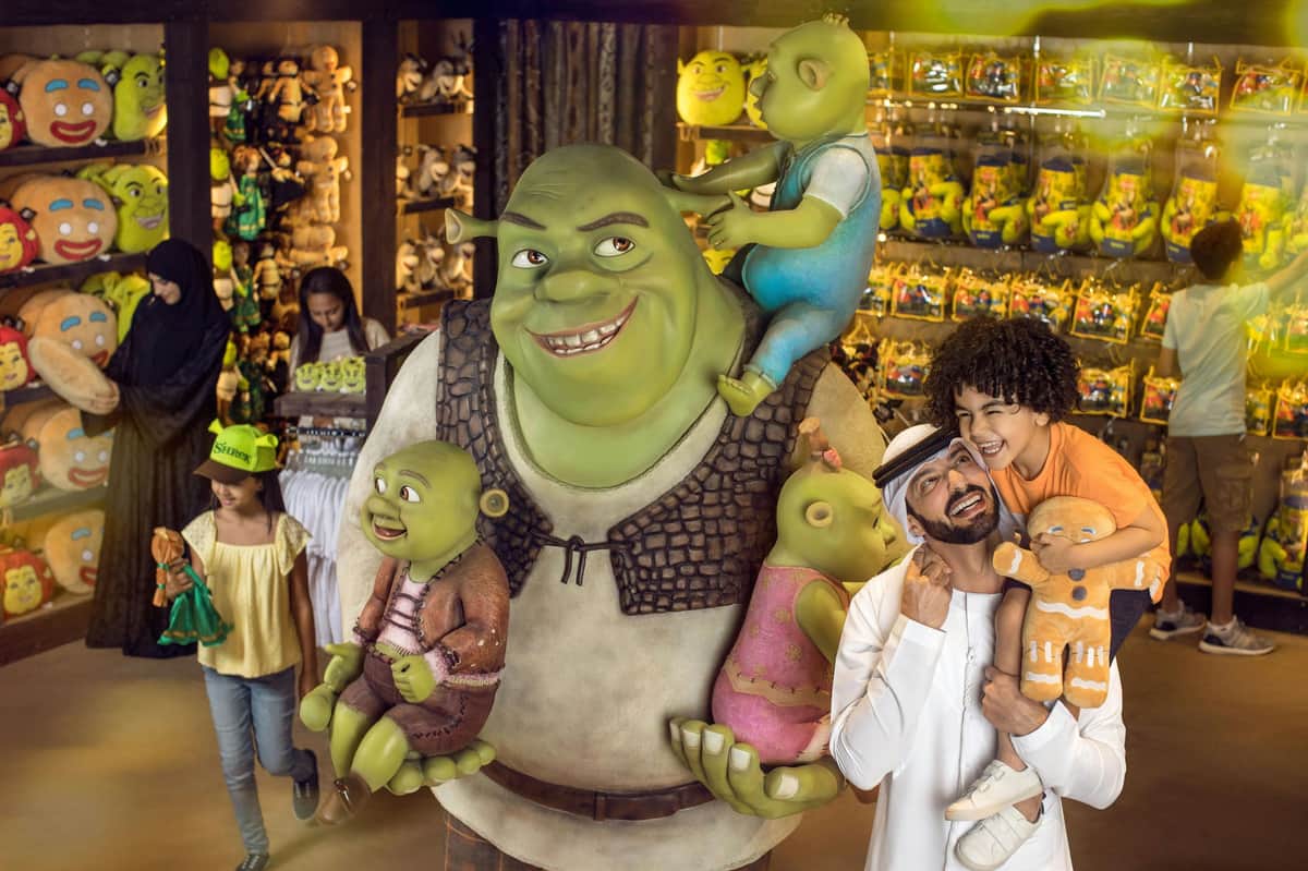 People posing with statues of Shrek and his family inside a brightly lit gift shop filled with Shrek-themed merchandise.