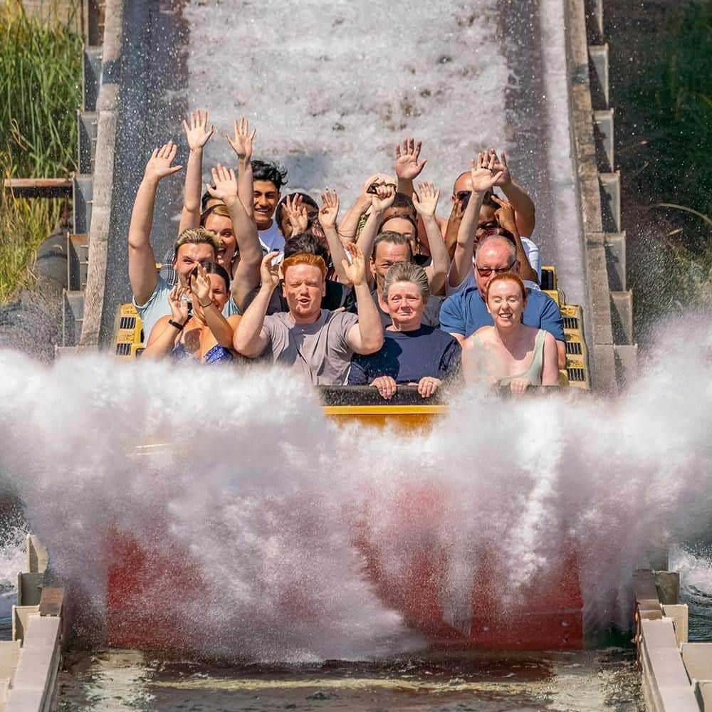 People on a log flume ride, raising their hands as water splashes up around them, creating waves on either side.