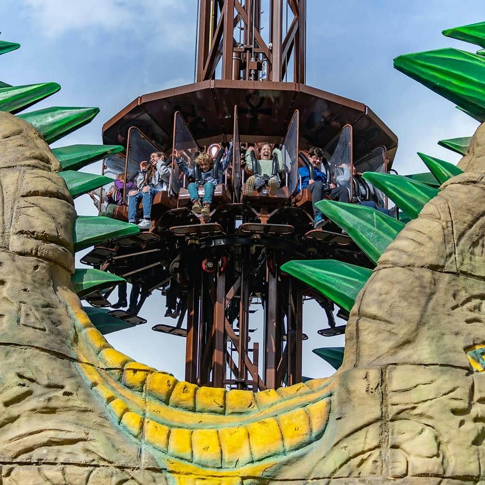 People ride a drop tower amusement ride, surrounded by large, decorative green spikes and stone structures under a blue sky.