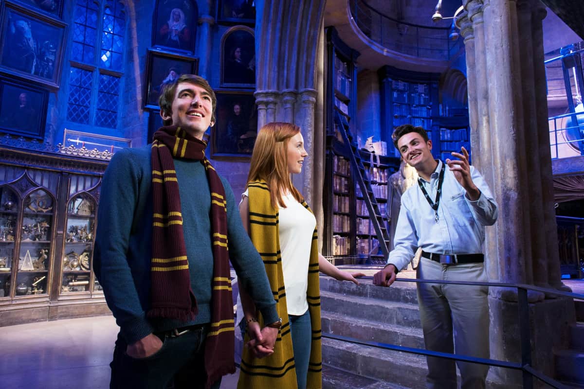 Harry Potter Warner Bros. Studio di Londra: Ingresso + Trasferimento dal British Museum Una guida spiega una mostra della biblioteca a una coppia che si tiene per mano e indossa sciarpe abbinate.