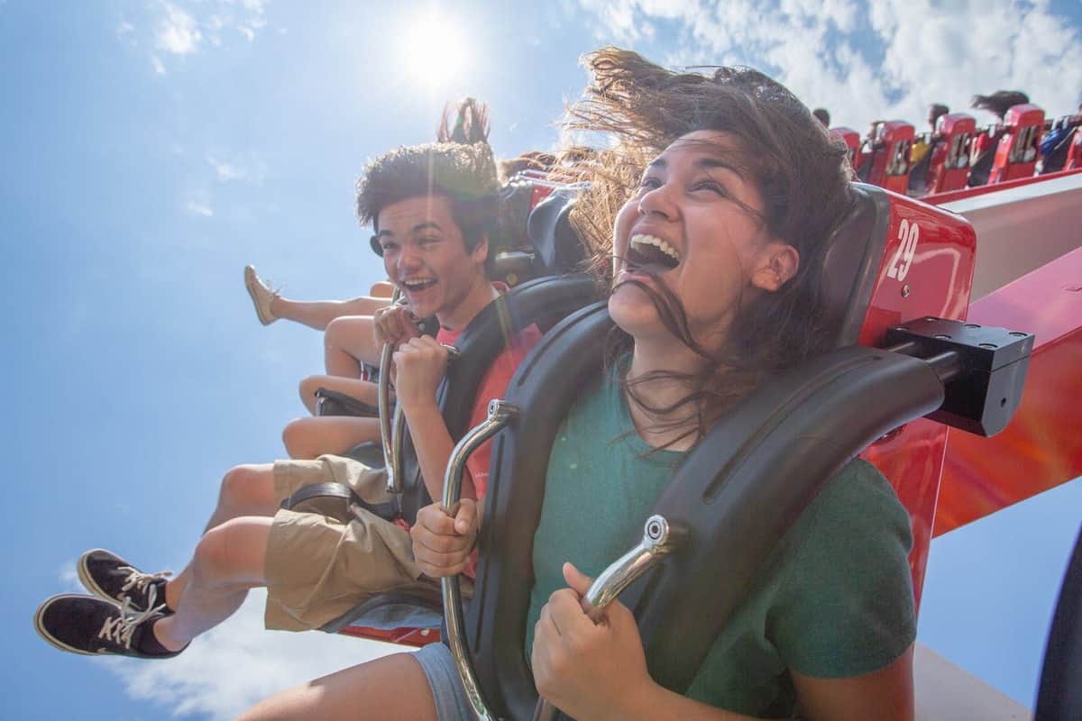 Roller coaster with red tracks and riders, surrounded by green trees, under a blue sky with scattered clouds.