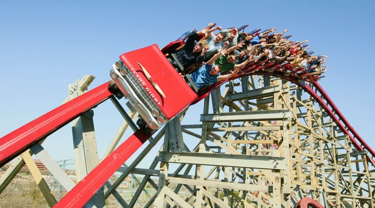 A red roller coaster shaped like a classic car with passengers leaning and holding their hands up, on a wooden structure.