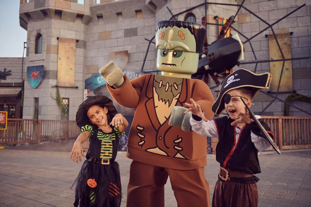 Child in a pirate costume and person in a large Frankenstein costume pose with smiles in front of a theme park backdrop.