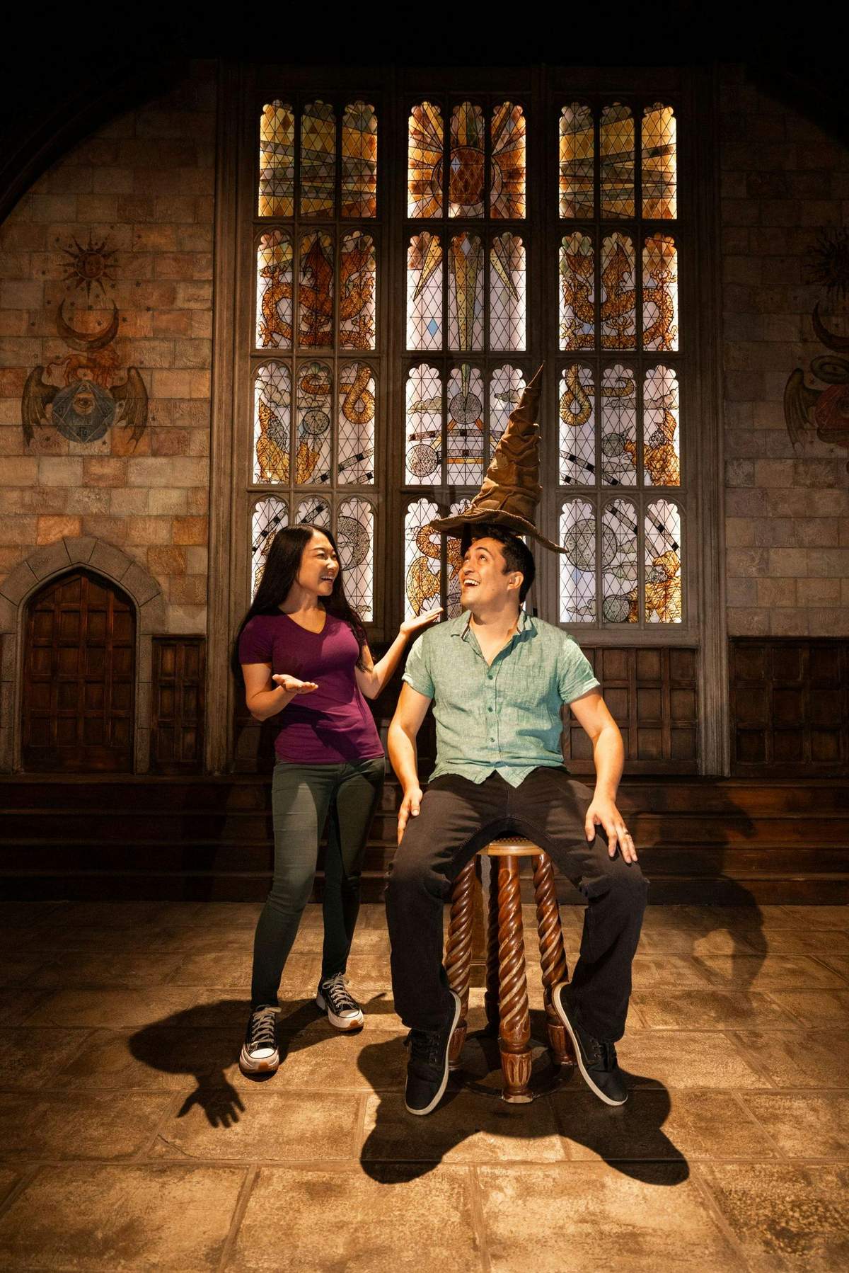 Two people in a stone room. One stands while the other sits on a stool wearing a wizard hat, in front of ornate stained glass windows.