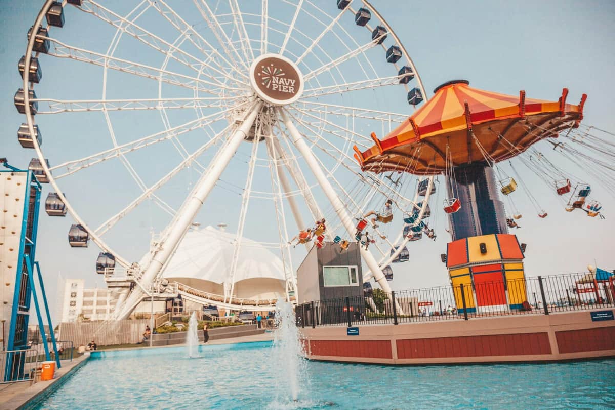 Ferris wheel and spinning carousel at Navy Pier with people riding, near a water fountain, under a clear sky.