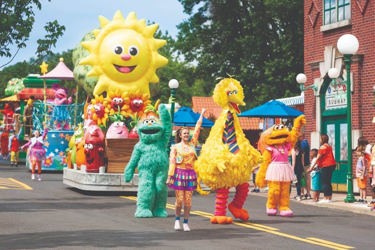 Colorful parade with people in Sesame Street costumes, including a large inflatable sun, smiling and waving.