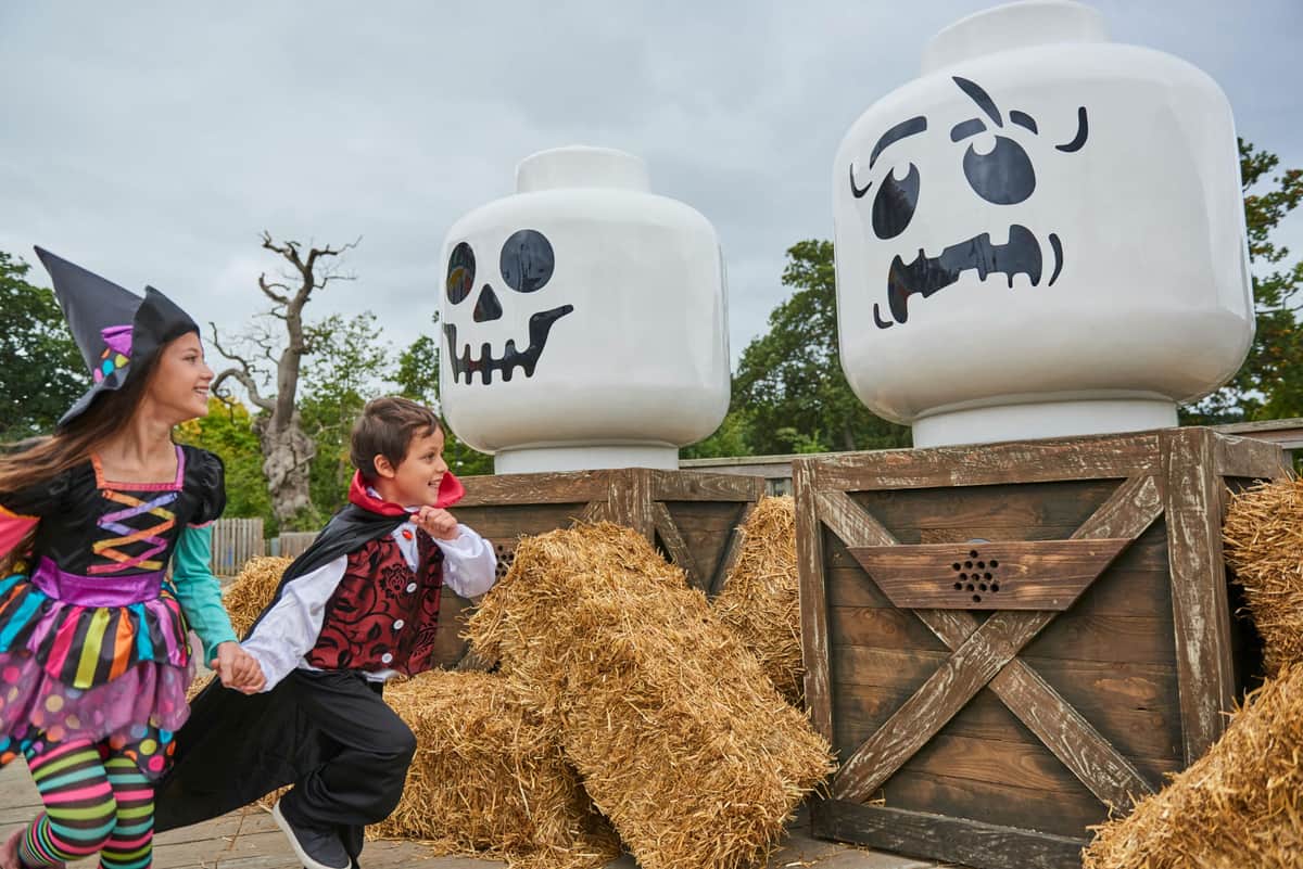 Two children in Halloween costumes run by large LEGO skull decorations and hay bales, with trees in the background.