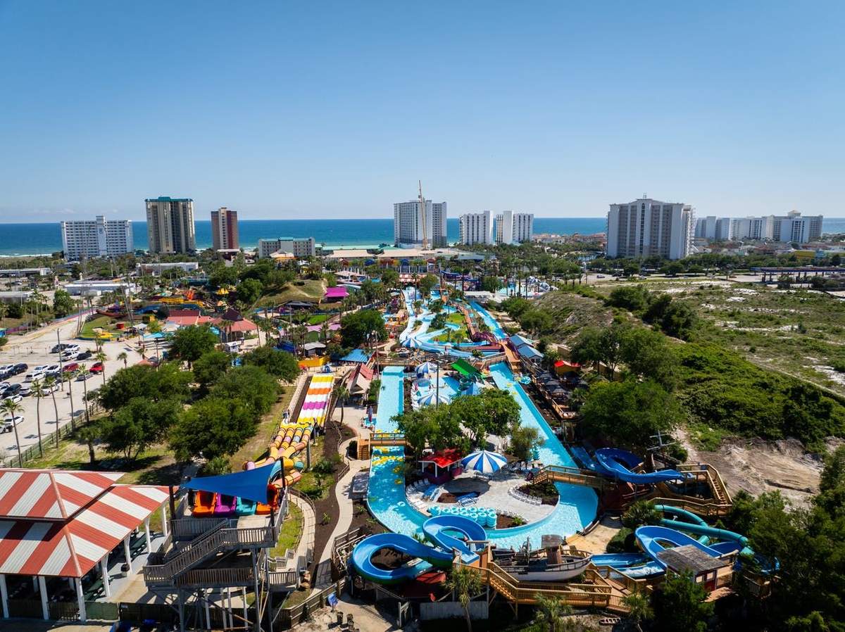 Aerial view of a water park with slides, pools, and colorful features, surrounded by trees and buildings, with the ocean beyond.
