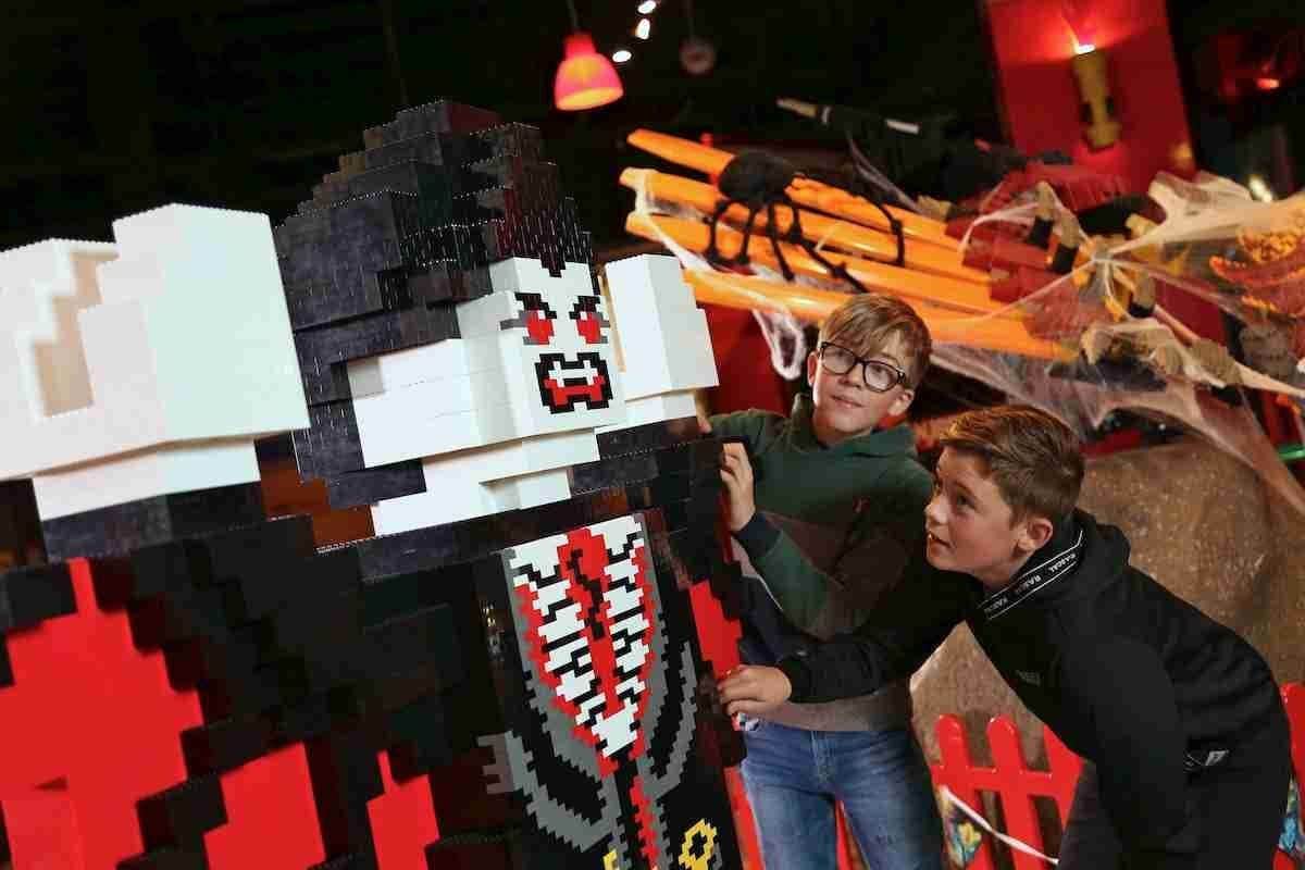 Two boys interact with a large LEGO vampire figure. Spiders and cobwebs decorate the background in a Halloween-themed setup.