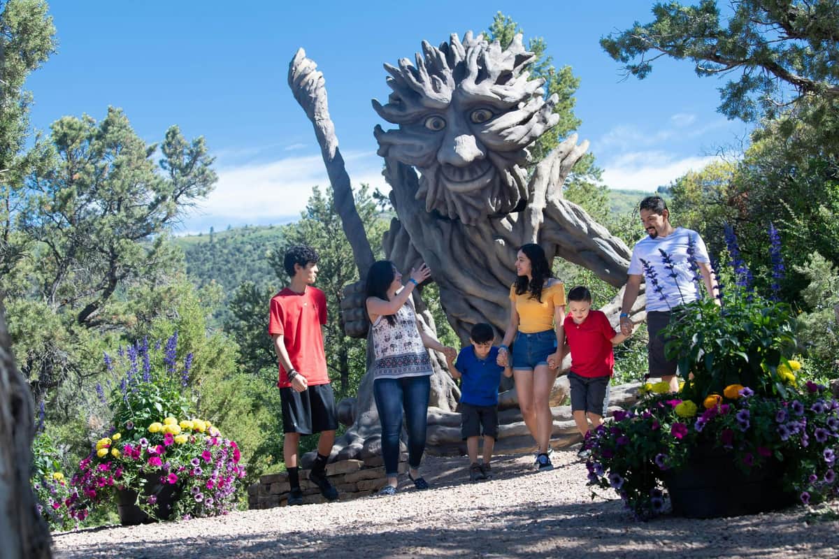 A family of six stands in front of a large tree-like sculpture with a smiling face, surrounded by greenery and flowers.