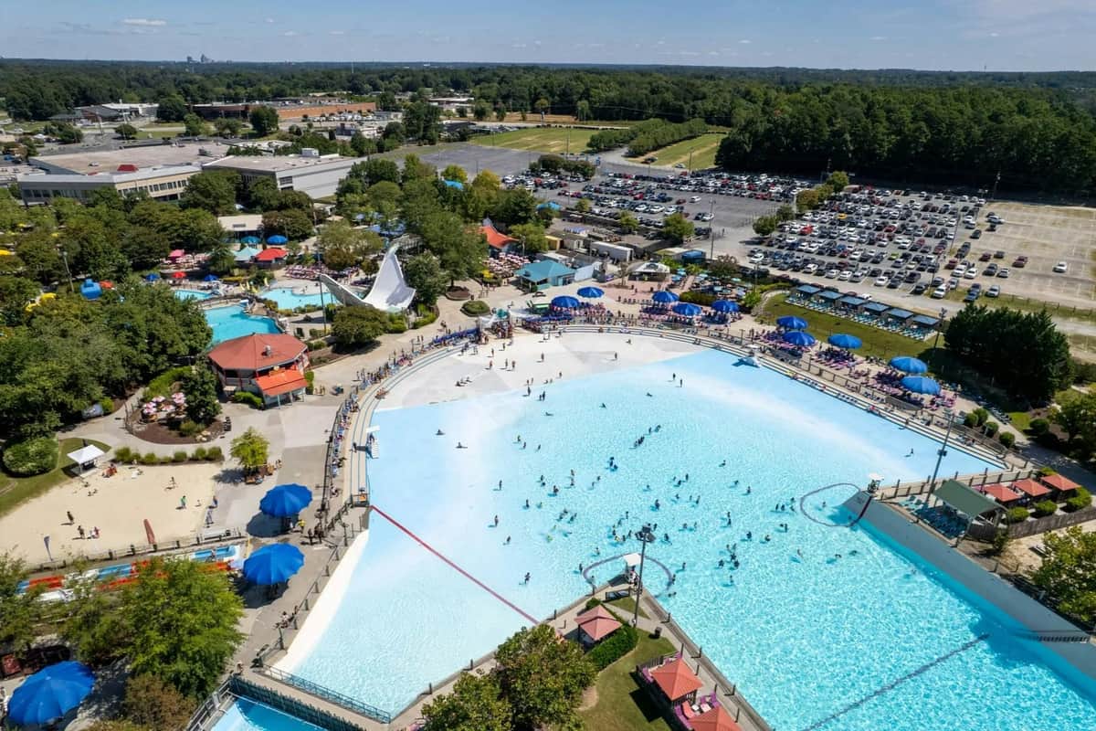 A large water park with a wave pool, water slides, and surrounding facilities, viewed from above on a sunny day.