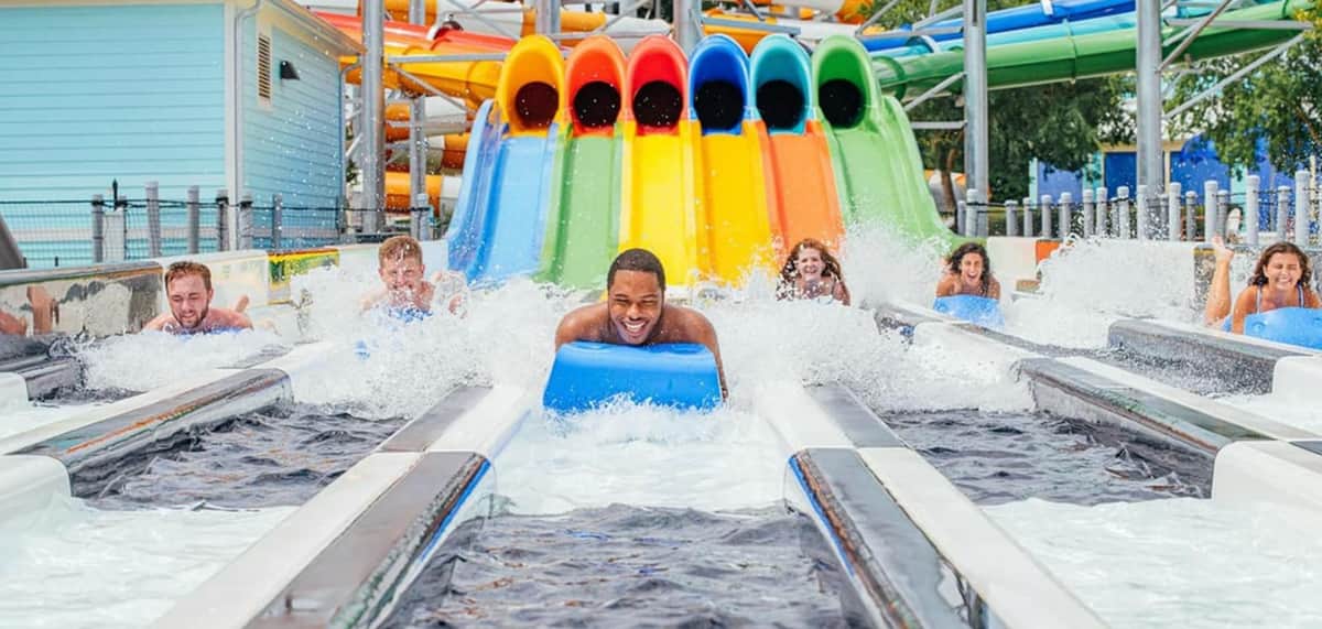 People racing down water slides on mats in a water park, splashing into the pool at the bottom with colorful slides in view.