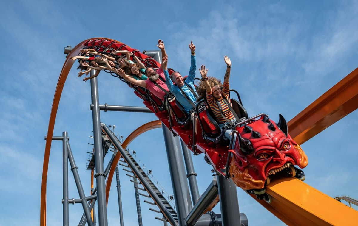 People on a rollercoaster with a dragon-themed front, raising their arms and smiling under a clear blue sky.