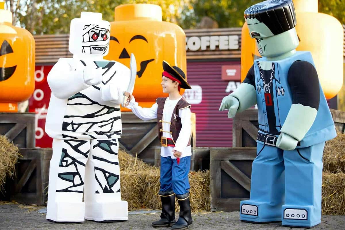 Child in a pirate costume interacts with life-sized LEGO mummy and Frankenstein figures in a Halloween-themed setting.