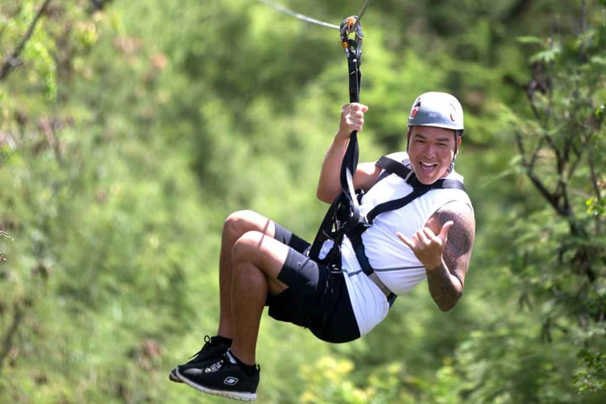 Person zip-lining through a lush green forest, wearing a helmet and safety harness, smiling and making a hand gesture.