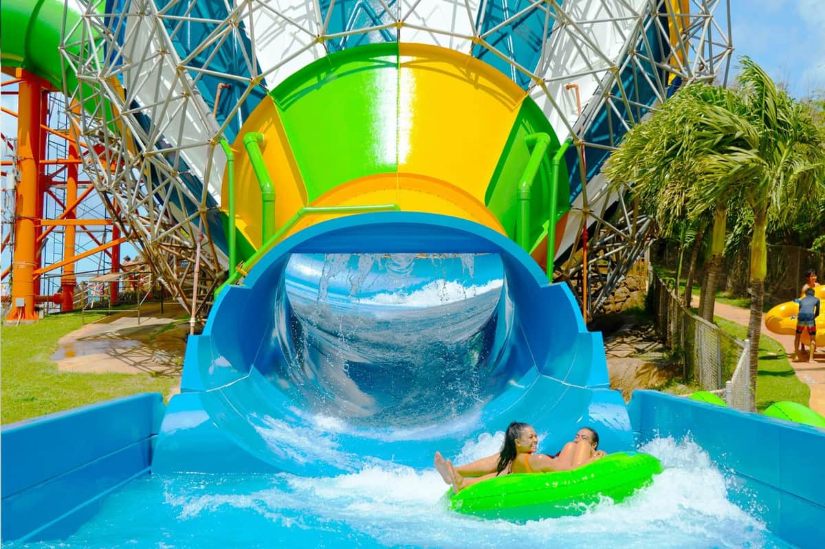 Parque del Cráter de Coral y Wet 'n' Wild Hawaii: Zip & Dip + Transporte desde Waikiki Dos personas en una balsa hinchable verde salen de un colorido tobogán acuático hacia una piscina, rodeadas de un entorno vibrante y soleado.