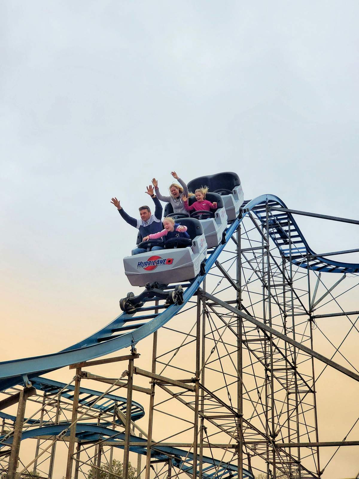 Four people riding a rollercoaster with raised hands and excited expressions as the car descends. Sky and coaster track in the background.