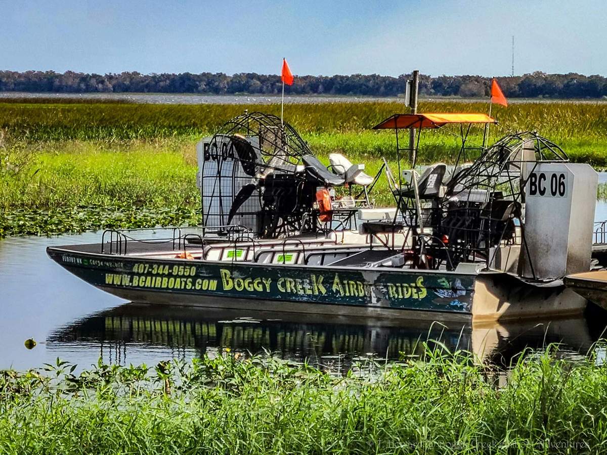 An airboat labeled "Boggy Creek Airboat Rides" is docked on a grassy waterway, equipped with seats, fan, and orange flags.