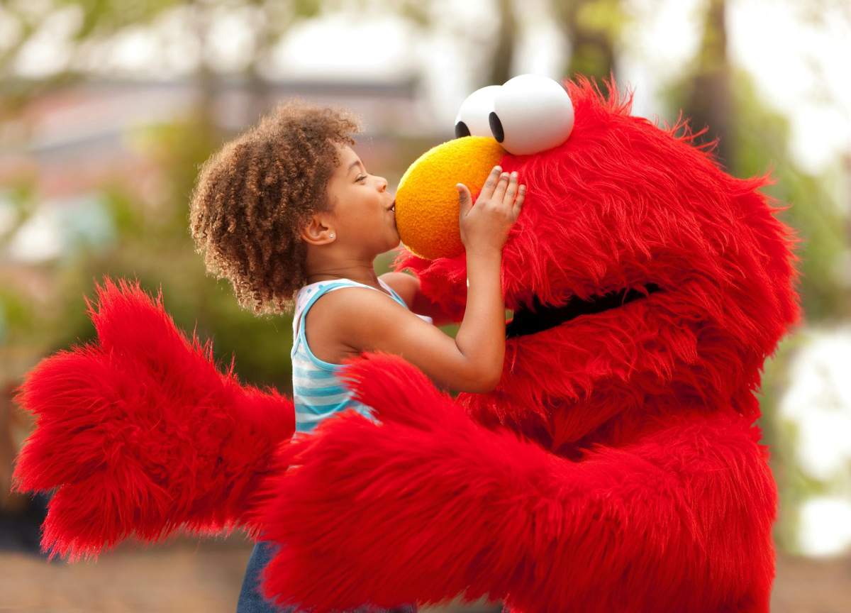 A child in a blue dress kisses a person in an Elmo costume outdoors, with greenery in the background.