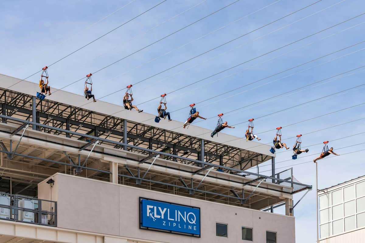People ziplining above a building with a sign that reads "Fly LINQ Zipline" and a clear blue sky in the background.
