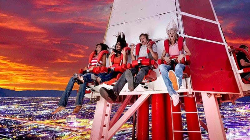 Four people in red harnesses on a thrill ride, showing expressions of excitement and fear, with a city skyline below at sunset.