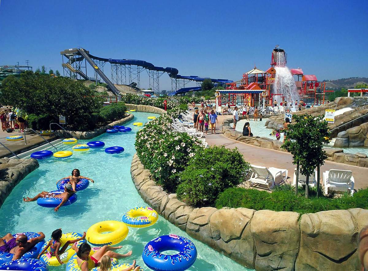 People float in a pool on inflatable tubes near a building with nautical decor and palm trees under a blue sky.
