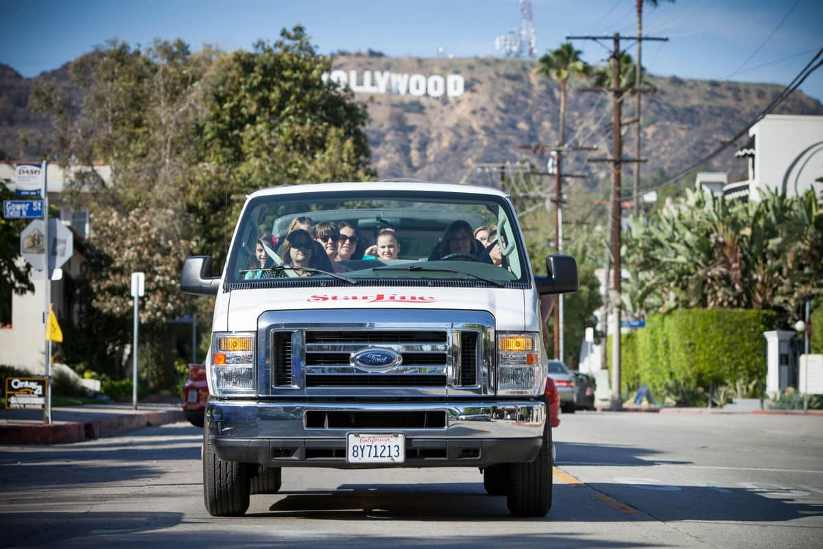 A van with passengers inside is driving on a street, with the Hollywood sign visible on a hill in the background.