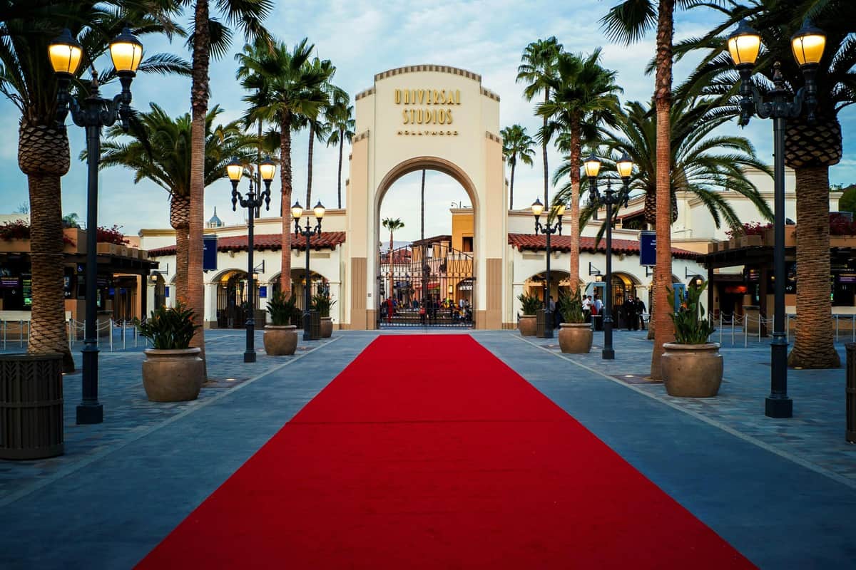 A red carpet leads to the Universal Studios Hollywood entrance arch, surrounded by palm trees and decorative lighting.