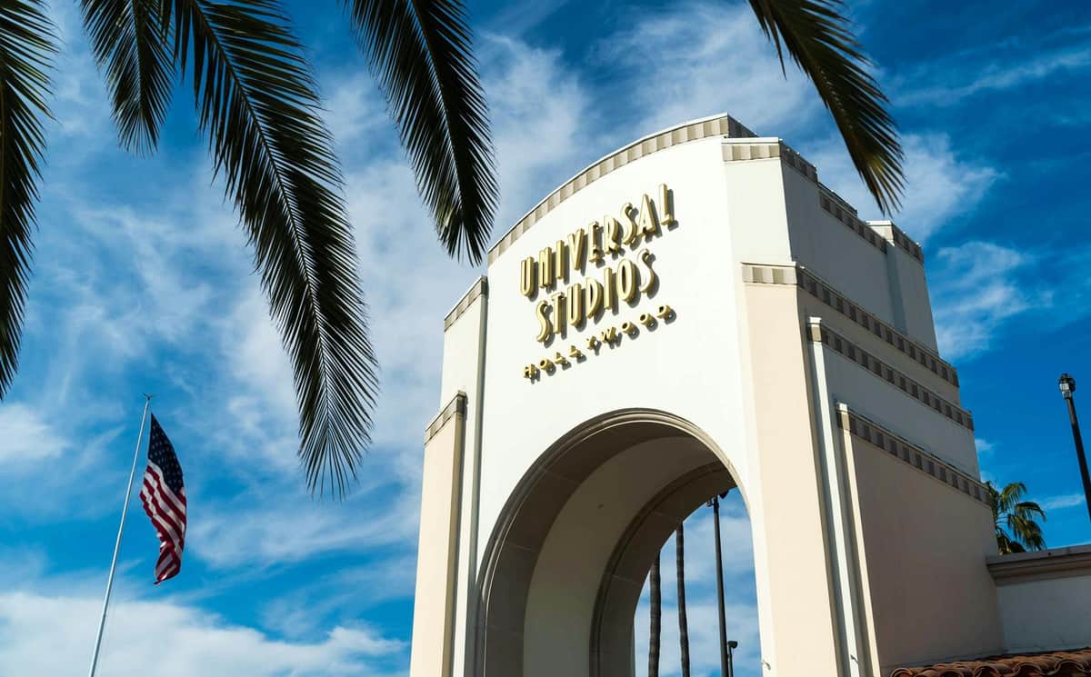 White archway with "Universal Studios Hollywood" signage, set against a blue sky. Palm tree leaves frame the top corners.